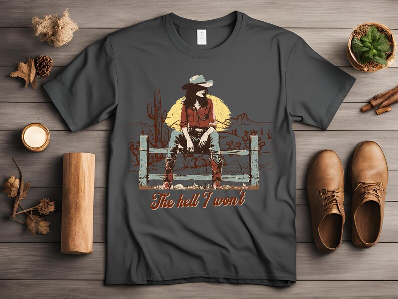 Op de afbeelding: Een donkergrijs T-shirt met een vintage illustratie van een vrouw die een cowboyhoed en laarzen draagt ​​en op een hek zit. De tekst "The hell I won't" is onder de illustratie gedrukt.