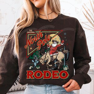 May include: Brown sweatshirt featuring a graphic of Santa Claus riding a reindeer in a rodeo setting. The design includes the text "The Original North Pole Rodeo" in red and green, with cacti and a full moon in the background.