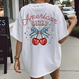 May include: White t-shirt with a red, white and blue cherry graphic and the text "American Girl" in a rope-like font.