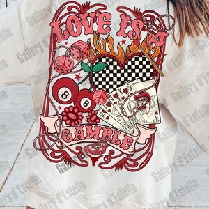 May include: White sweatshirt with a graphic design featuring the words "LOVE IS A GAMBLE" in pink, along with dice, playing cards, cherries, and an eight ball. The design has a vintage, retro aesthetic.