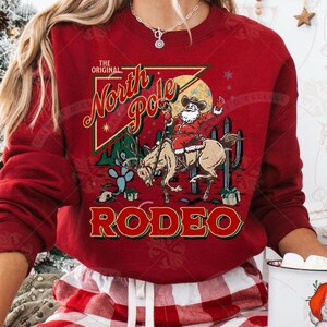 May include: A red sweatshirt with a graphic featuring Santa Claus riding a horse in a rodeo setting. The text reads "The Original North Pole Rodeo" in a vintage style. Cactus and gift boxes are also in the design.