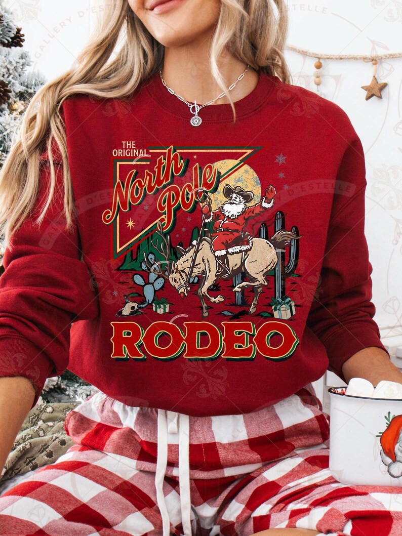 May include: A red sweatshirt with a graphic featuring Santa Claus riding a horse in a rodeo setting. The text reads "The Original North Pole Rodeo" in a vintage style. Cactus and gift boxes are also in the design.