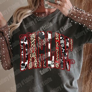 May include: Dark gray t-shirt with the word "Darlin" in a patchwork design. The letters feature cow print, leopard print, and black and white patterns, outlined in red. The sleeves are sheer with white pearl accents.
