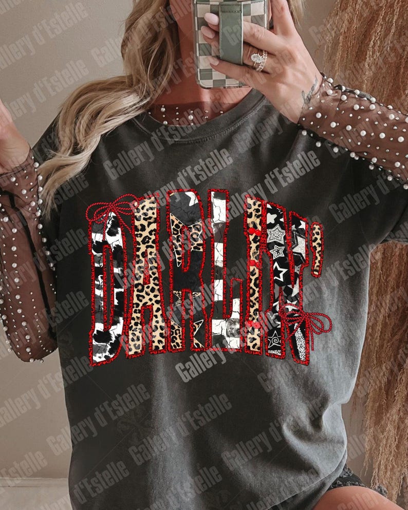May include: Dark gray t-shirt with the word "Darlin" in a patchwork design. The letters feature cow print, leopard print, and black and white patterns, outlined in red. The sleeves are sheer with white pearl accents.