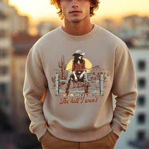 Op de afbeelding: Een beige sweatshirt met een grafische print van een vrouw die een cowboyhoed draagt en op een hek zit. De tekst "The hell I won't" is onder de print gedrukt.