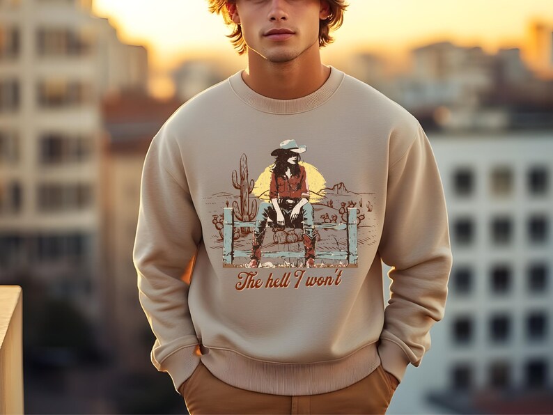 Op de afbeelding: Een beige sweatshirt met een grafische print van een vrouw die een cowboyhoed draagt en op een hek zit. De tekst "The hell I won't" is onder de print gedrukt.