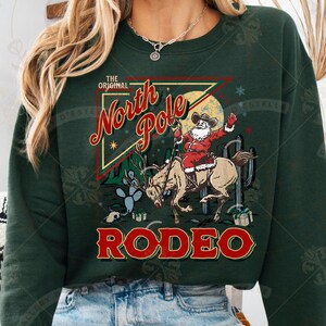 May include: Dark green sweatshirt with a graphic of Santa Claus riding a bucking bronco in a rodeo setting. The design includes the words "The Original North Pole Rodeo" in red and yellow lettering, with cacti and a crescent moon.