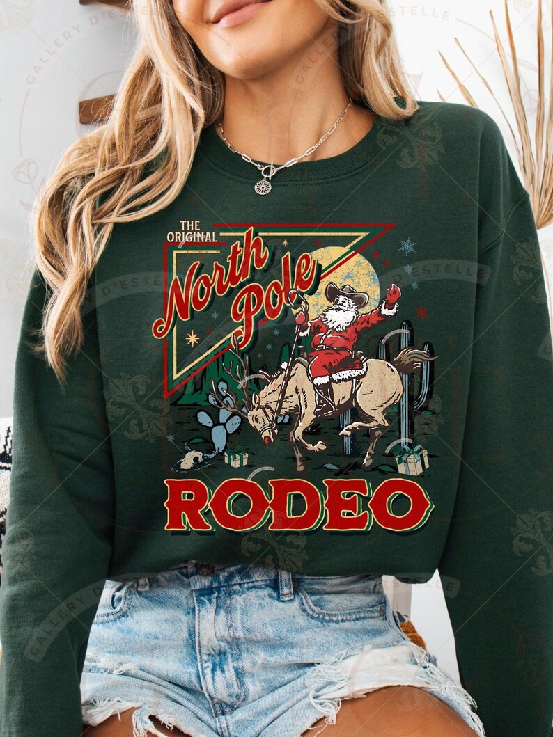 May include: Dark green sweatshirt with a graphic of Santa Claus riding a bucking bronco in a rodeo setting. The design includes the words "The Original North Pole Rodeo" in red and yellow lettering, with cacti and a crescent moon.