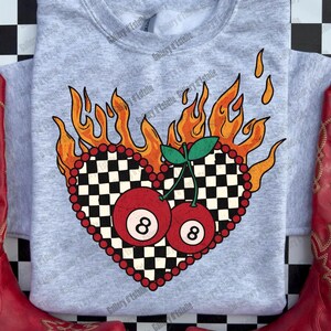 May include: A light gray sweatshirt featuring a heart-shaped design with a checkered pattern and two red 8-ball cherries. The heart is surrounded by orange flames and red beads. Red cowboy boots are on either side of the sweatshirt.