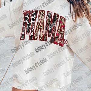 May include: Cream-colored sweatshirt with the word "MAMA" in a bold, decorative font. The letters feature a mix of patterns, including cow print, leopard print, and floral designs, outlined in red. A stylish and comfortable top.