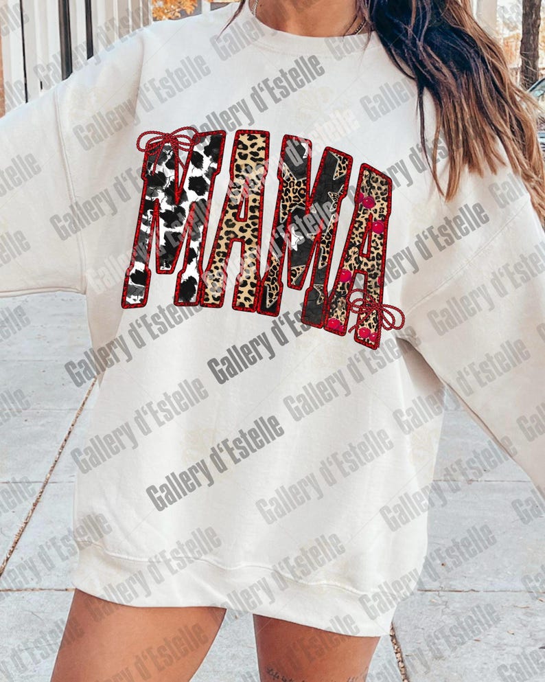 May include: Cream-colored sweatshirt with the word "MAMA" in a bold, decorative font. The letters feature a mix of patterns, including cow print, leopard print, and floral designs, outlined in red. A stylish and comfortable top.
