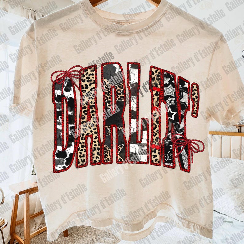 May include: A beige t-shirt with the word "DARLIN" in a patchwork design. The letters feature cow print, leopard print, and star patterns, outlined in red. The shirt is hanging on a wooden hanger.