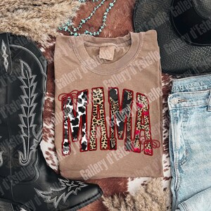 May include: A tan-colored t-shirt with the word "MAMA" in a bold, decorative font. The letters feature a mix of animal print patterns, including cow print and leopard print, outlined in red. The shirt is styled with a cowboy hat, boots, and jeans.