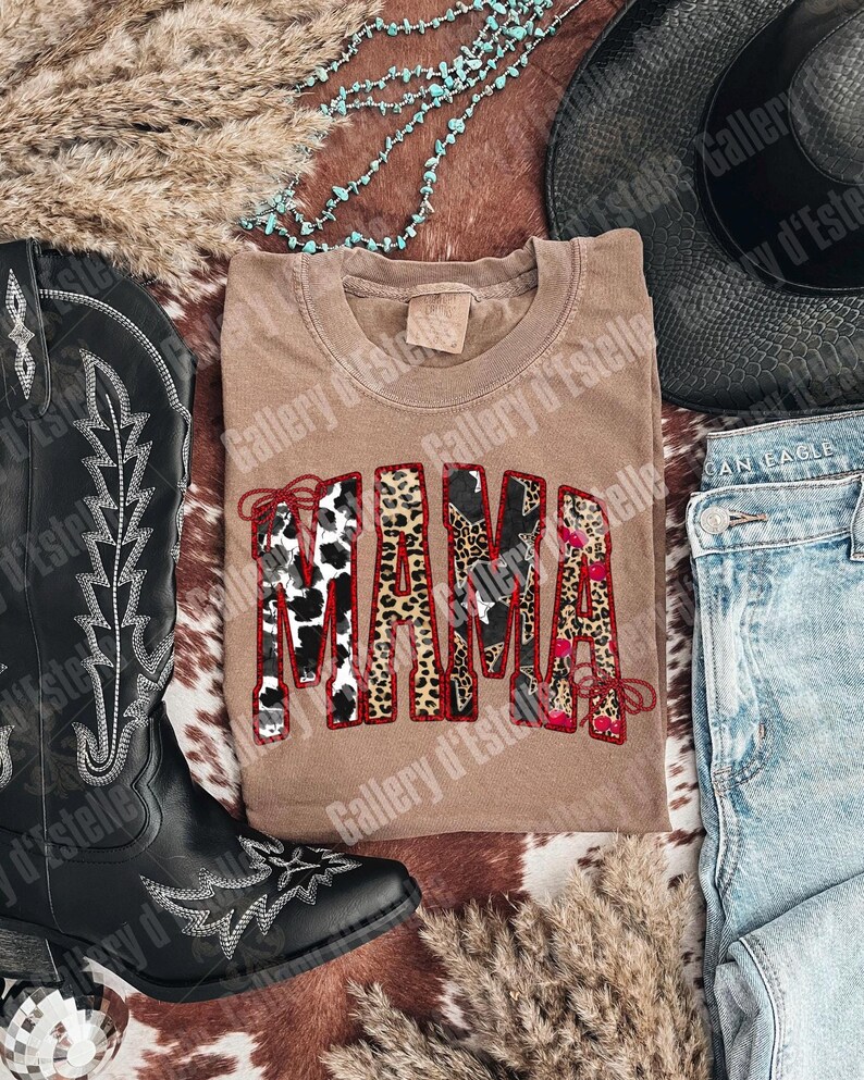 May include: A tan-colored t-shirt with the word "MAMA" in a bold, decorative font. The letters feature a mix of animal print patterns, including cow print and leopard print, outlined in red. The shirt is styled with a cowboy hat, boots, and jeans.