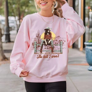 Op de afbeelding: Roze sweatshirt met een grafische print van een vrouw in een cowboyhoed die op een hek zit met de tekst "The hell I won't".