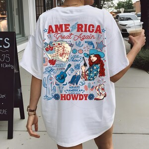 以下が含まれることがあります： アメリカ国旗をモチーフにしたグラフィックプリントが施された白いTシャツ。カウボーイハット、星、花火、そして「AMERICA Great Again... HOWDY」の文字がデザインされています。
