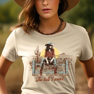 Op de afbeelding: Een beige T-shirt met een afbeelding van een vrouw in een cowboyhoed die op een hek zit. De afbeelding bevat cactussen en de tekst "The hell I won't".