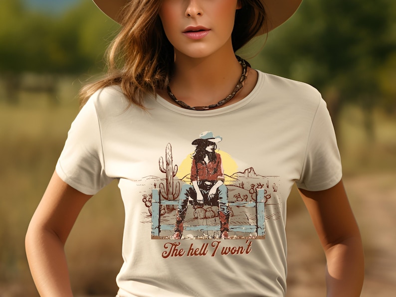 Op de afbeelding: Een beige T-shirt met een afbeelding van een vrouw in een cowboyhoed die op een hek zit. De afbeelding bevat cactussen en de tekst "The hell I won't".