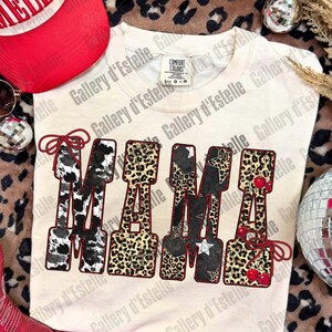 May include: Cream-colored Comfort Colors t-shirt with the word "MAMA" in a bold, decorative font. The letters feature cow print, leopard print, stars, and cherries. A red and white baseball cap and red boots are visible.