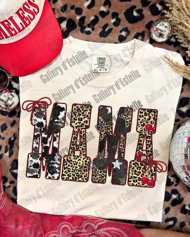 May include: Cream-colored Comfort Colors t-shirt with the word "MAMA" in a bold, decorative font. The letters feature cow print, leopard print, stars, and cherries. A red and white baseball cap and red boots are visible.