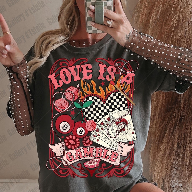 May include: Dark gray t-shirt with a graphic design featuring the text "LOVE IS A GAMBLE". The design includes dice, cherries, playing cards, and a checkered heart. The shirt has a vintage, distressed look.
