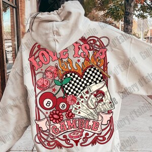 May include: Cream-colored hoodie with a large graphic on the back. The graphic features the words "Love is a Gamble" in pink, along with dice, playing cards, cherries, and an 8-ball. The design has a retro, vintage aesthetic.