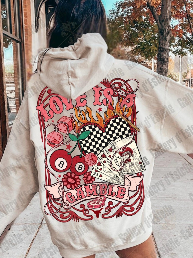 May include: Cream-colored hoodie with a large graphic on the back. The graphic features the words "Love is a Gamble" in pink, along with dice, playing cards, cherries, and an 8-ball. The design has a retro, vintage aesthetic.