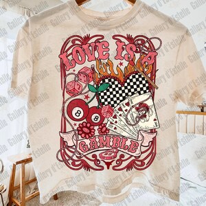 May include: Cream-colored t-shirt featuring a retro design with the words "LOVE IS GAMBLE" in pink. The graphic includes dice, playing cards, cherries, and a checkered heart, evoking a vintage casino theme.