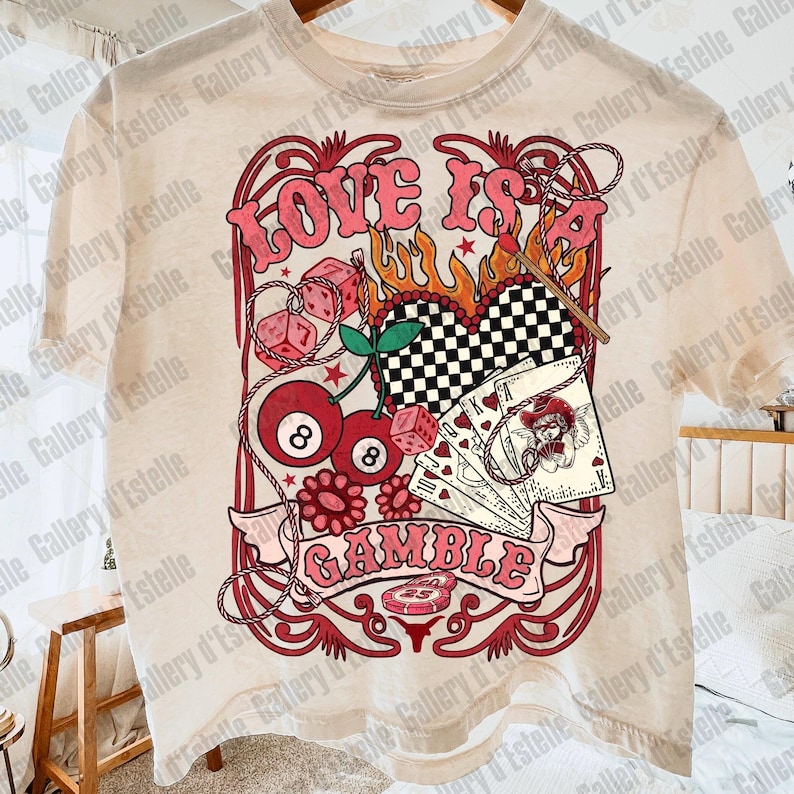 May include: Cream-colored t-shirt featuring a retro design with the words "LOVE IS GAMBLE" in pink. The graphic includes dice, playing cards, cherries, and a checkered heart, evoking a vintage casino theme.