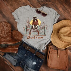 Op de afbeelding: Een grijs T-shirt met een afbeelding van een vrouw in een cowboyhoed en jeans die op een hek zit. De tekst "The hell I won't" is onder de afbeelding gedrukt. Het T-shirt wordt gecombineerd met een spijkerbroekje, bruine cowboylaarzen, een bruine leren tas en een strohoed.