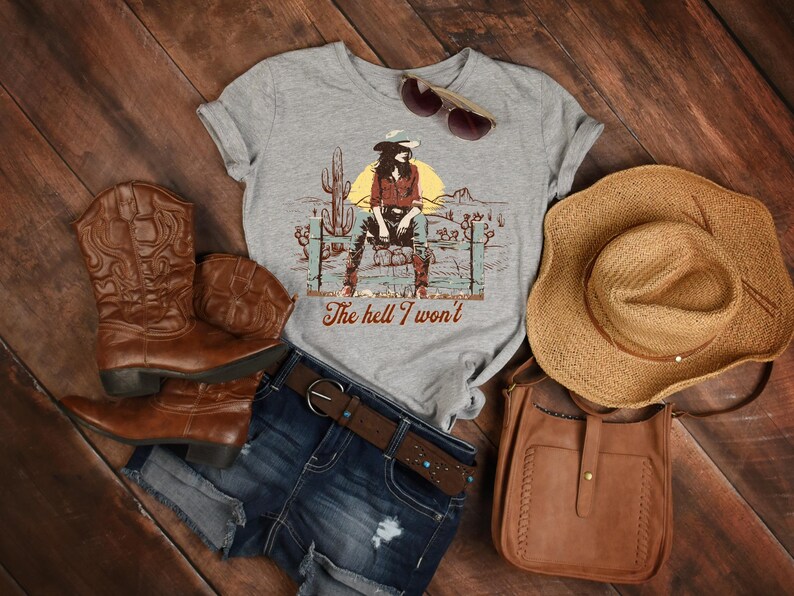 Op de afbeelding: Een grijs T-shirt met een afbeelding van een vrouw in een cowboyhoed en jeans die op een hek zit. De tekst "The hell I won't" is onder de afbeelding gedrukt. Het T-shirt wordt gecombineerd met een spijkerbroekje, bruine cowboylaarzen, een bruine leren tas en een strohoed.