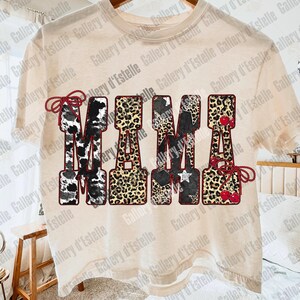 May include: Cream-colored t-shirt with the word "MAMA" in a bold, decorative font. The letters feature various patterns, including cow print, leopard print, stars, and cherries. The design is outlined in a dark red.