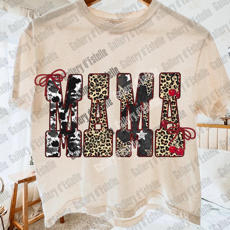 May include: Cream-colored t-shirt with the word "MAMA" in a bold, decorative font. The letters feature various patterns, including cow print, leopard print, stars, and cherries. The design is outlined in a dark red.