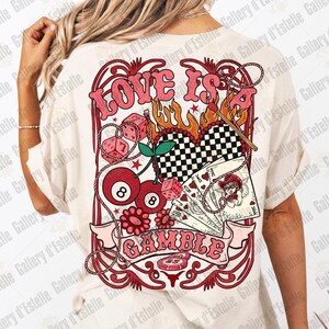 May include: Cream-colored t-shirt with a graphic design featuring the text "LOVE IS A GAMBLE". The design includes dice, playing cards, cherries, and a checkered heart, all in shades of red and pink.