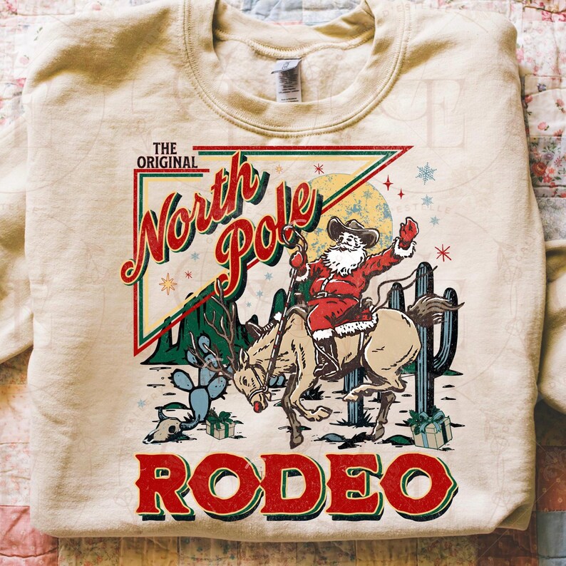 May include: A beige sweatshirt featuring a graphic of Santa Claus riding a horse in a rodeo setting. The design includes the text "THE ORIGINAL North Pole RODEO" in red and green, with cacti and stars.
