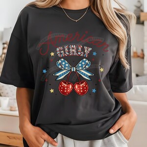 May include: A black t-shirt featuring a graphic of a red, white and blue bow with cherries and stars. The text "American Girl" is written above the bow.