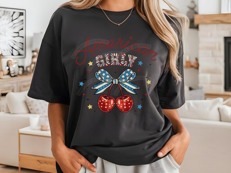 May include: A black t-shirt featuring a graphic of a red, white and blue bow with cherries and stars. The text "American Girl" is written above the bow.
