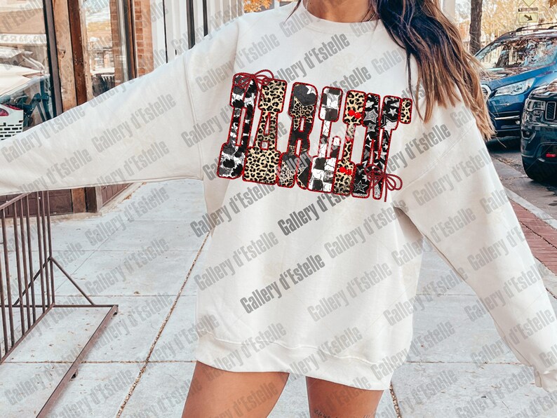 May include: Cream-colored sweatshirt with a graphic design that spells out "Spirit" using various patterns, including leopard print, stars, and cowhide. The word is accented with a red bow. The sweatshirt is a casual, comfortable style.