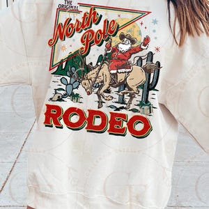 May include: Cream-colored sweatshirt featuring a festive graphic. The design includes Santa Claus riding a reindeer in a rodeo setting, with the words "North Pole Rodeo" in red and green. Cactus and snowflakes complete the holiday theme.
