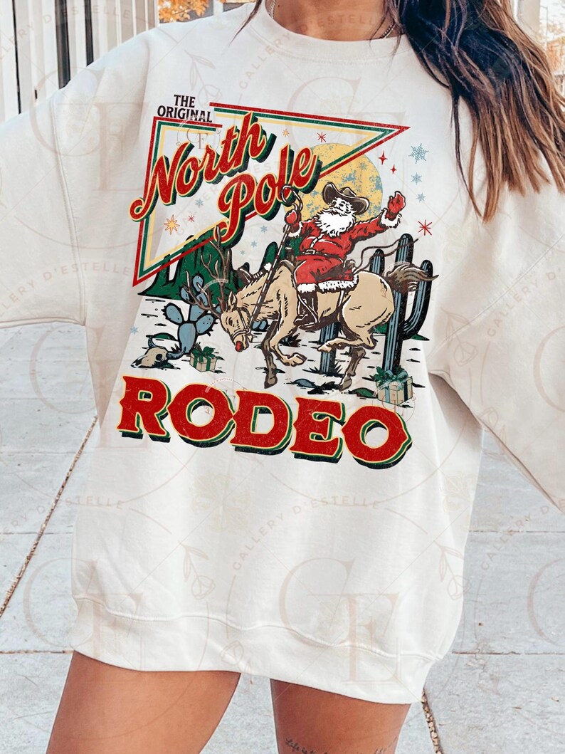 May include: Cream-colored sweatshirt featuring a festive graphic. The design includes Santa Claus riding a reindeer in a rodeo setting, with the words "North Pole Rodeo" in red and green. Cactus and snowflakes complete the holiday theme.