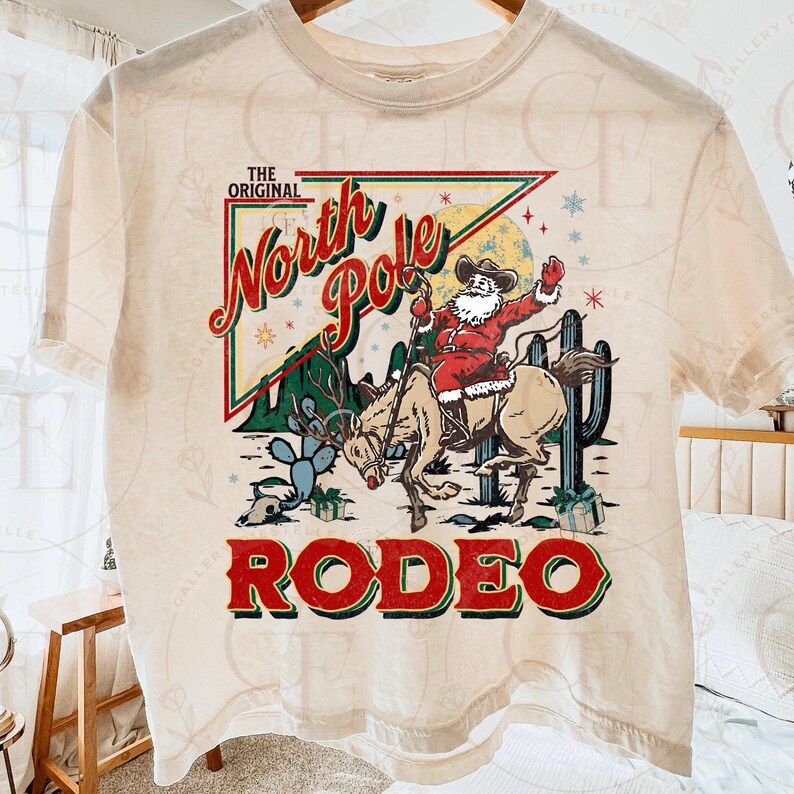 May include: Cream-colored t-shirt featuring a graphic of Santa Claus riding a reindeer at a rodeo. The design includes the text "THE ORIGINAL North Pole RODEO" in red and green, with cacti and a desert landscape.