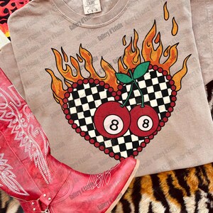 May include: A tan t-shirt featuring a heart-shaped design with a black and white checkered pattern, surrounded by flames and red beads. Two red 8-ball cherries are in the center. A red cowboy boot is in the foreground.