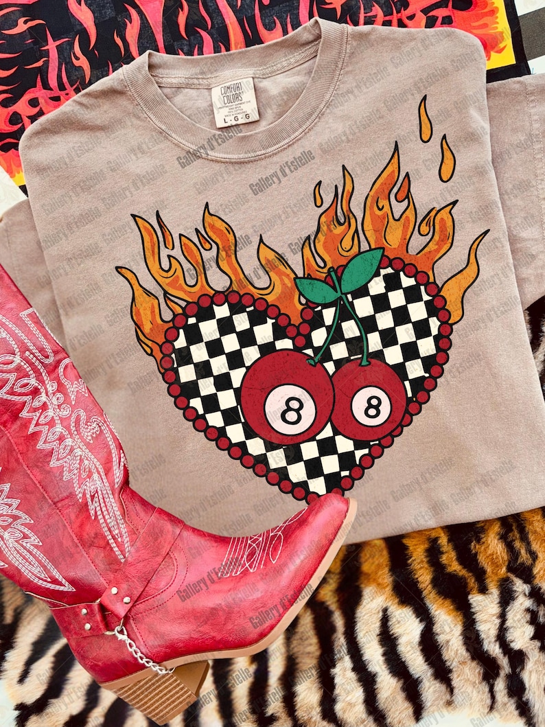 May include: A tan t-shirt featuring a heart-shaped design with a black and white checkered pattern, surrounded by flames and red beads. Two red 8-ball cherries are in the center. A red cowboy boot is in the foreground.