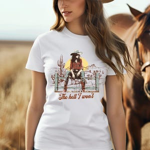 Op de afbeelding: Wit T-shirt met een afbeelding van een vrouw met een cowboyhoed die op een hek zit. De tekst "The hell I won't" staat onder de afbeelding.