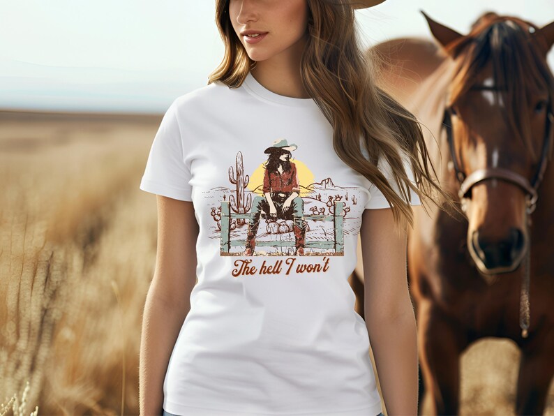 Op de afbeelding: Wit T-shirt met een afbeelding van een vrouw met een cowboyhoed die op een hek zit. De tekst "The hell I won't" staat onder de afbeelding.