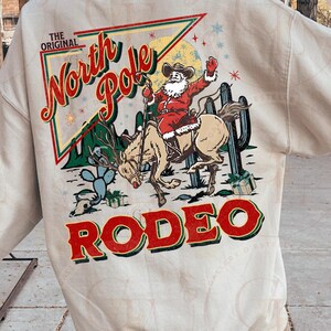 May include: Beige sweatshirt with a graphic of Santa Claus riding a horse in a rodeo setting. The design includes the words "The Original North Pole Rodeo" in red and green, with cactus and gift box illustrations.