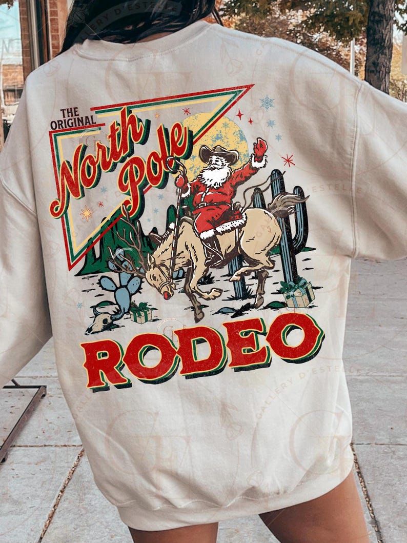 May include: Beige sweatshirt with a graphic of Santa Claus riding a horse in a rodeo setting. The design includes the words "The Original North Pole Rodeo" in red and green, with cactus and gift box illustrations.