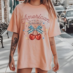 May include: A pink t-shirt with the text "American Cherry" in a glittery font. The shirt features a cherry with a red, white and blue bow.