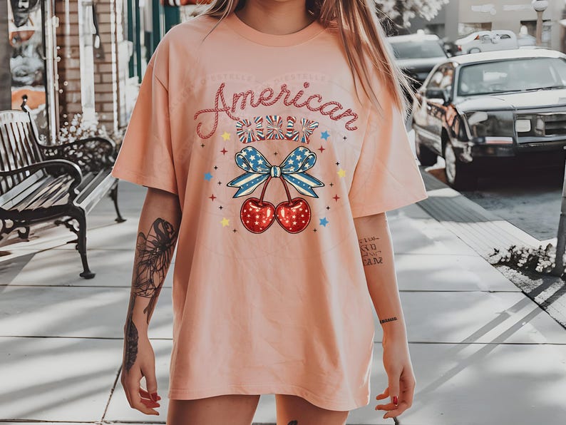 May include: A pink t-shirt with the text "American Cherry" in a glittery font. The shirt features a cherry with a red, white and blue bow.