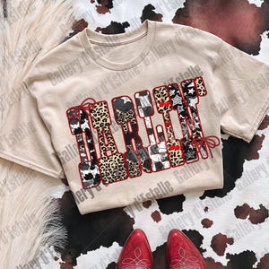 May include: A beige t-shirt with the word "TAKE IT" in a patchwork design featuring cow print, leopard print, stars, and red accents. Red cowboy boots are at the bottom of the image, set against a cowhide pattern.